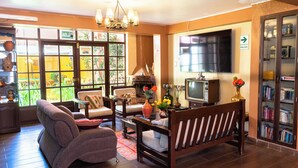 Una sala de estar acogedora con sofá, sillón y televisor. Se observa una estantería y un ventanal con vistas al exterior.