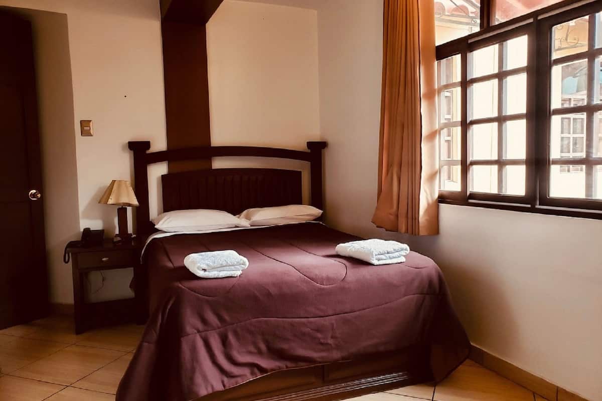 A neatly made bed with a dark headboard, a bedside table with a lamp, and a window with curtains.