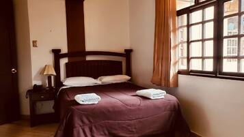 A neatly made bed with a dark headboard, a bedside table with a lamp, and a window with curtains.