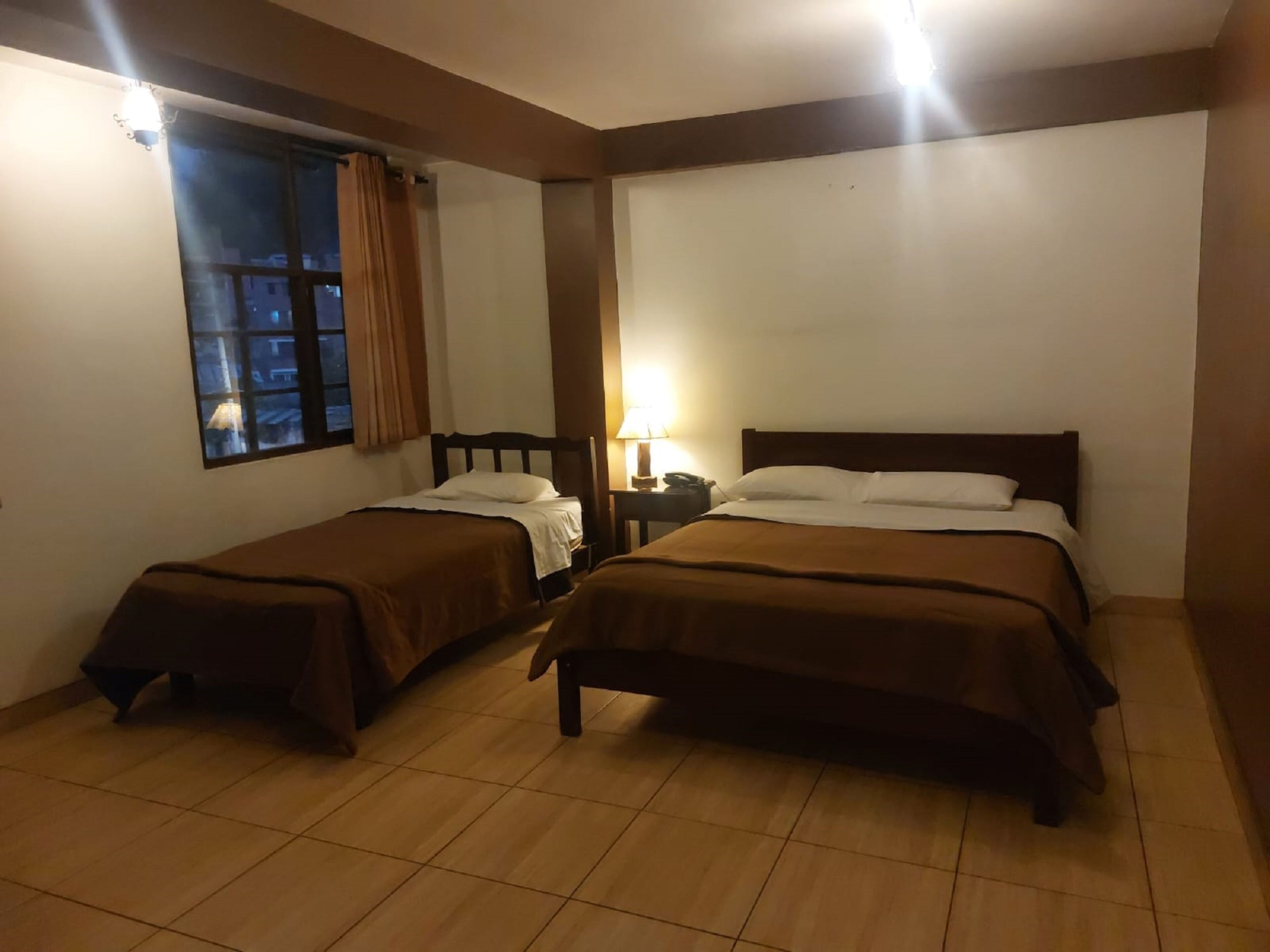 A hotel room with two beds, a window with curtains, a nightstand with a lamp, and a tiled floor.