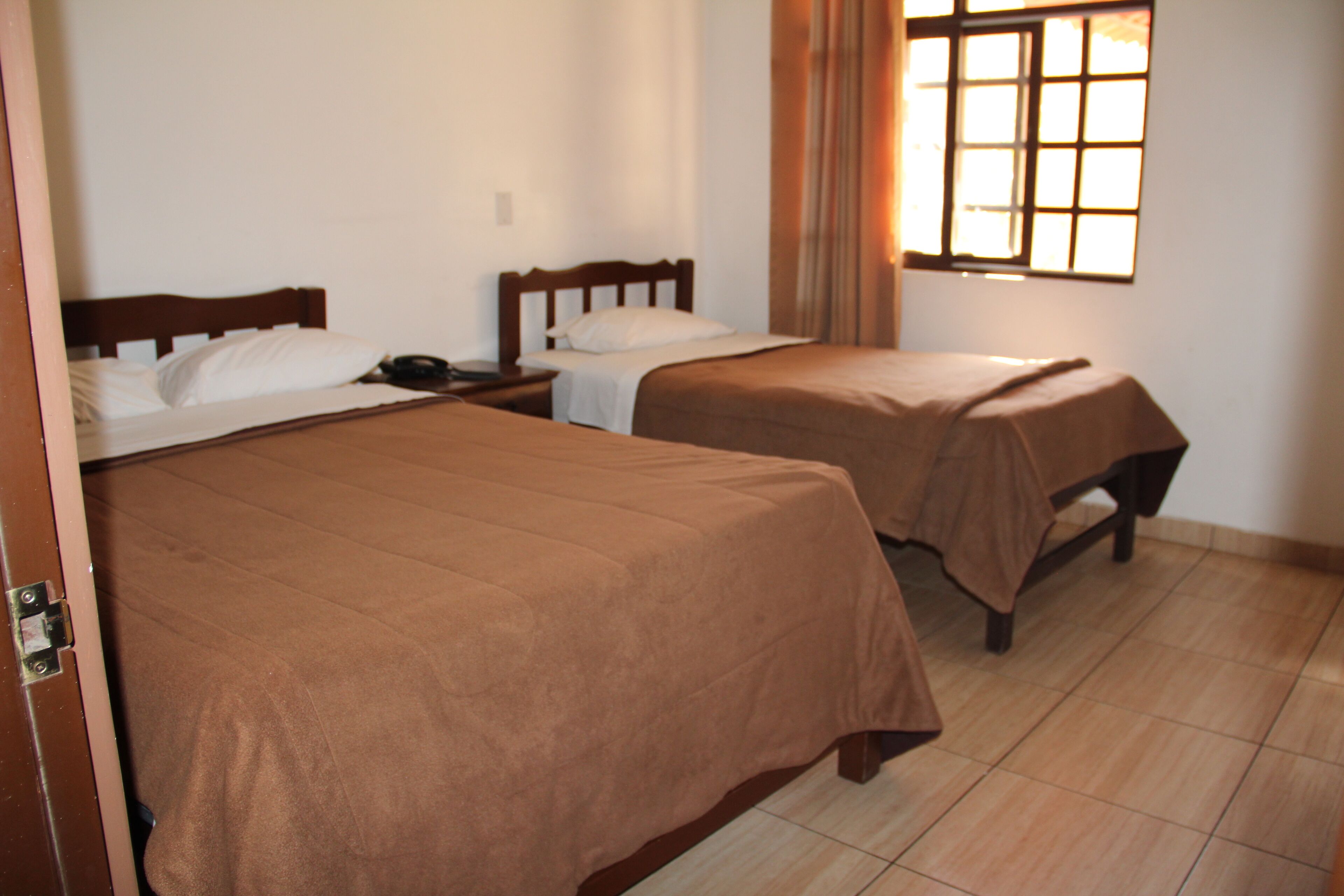 A room with two beds, each with a wooden headboard and a brown bedspread. There is a window with a curtain on the right side, and the room has tiled flooring.