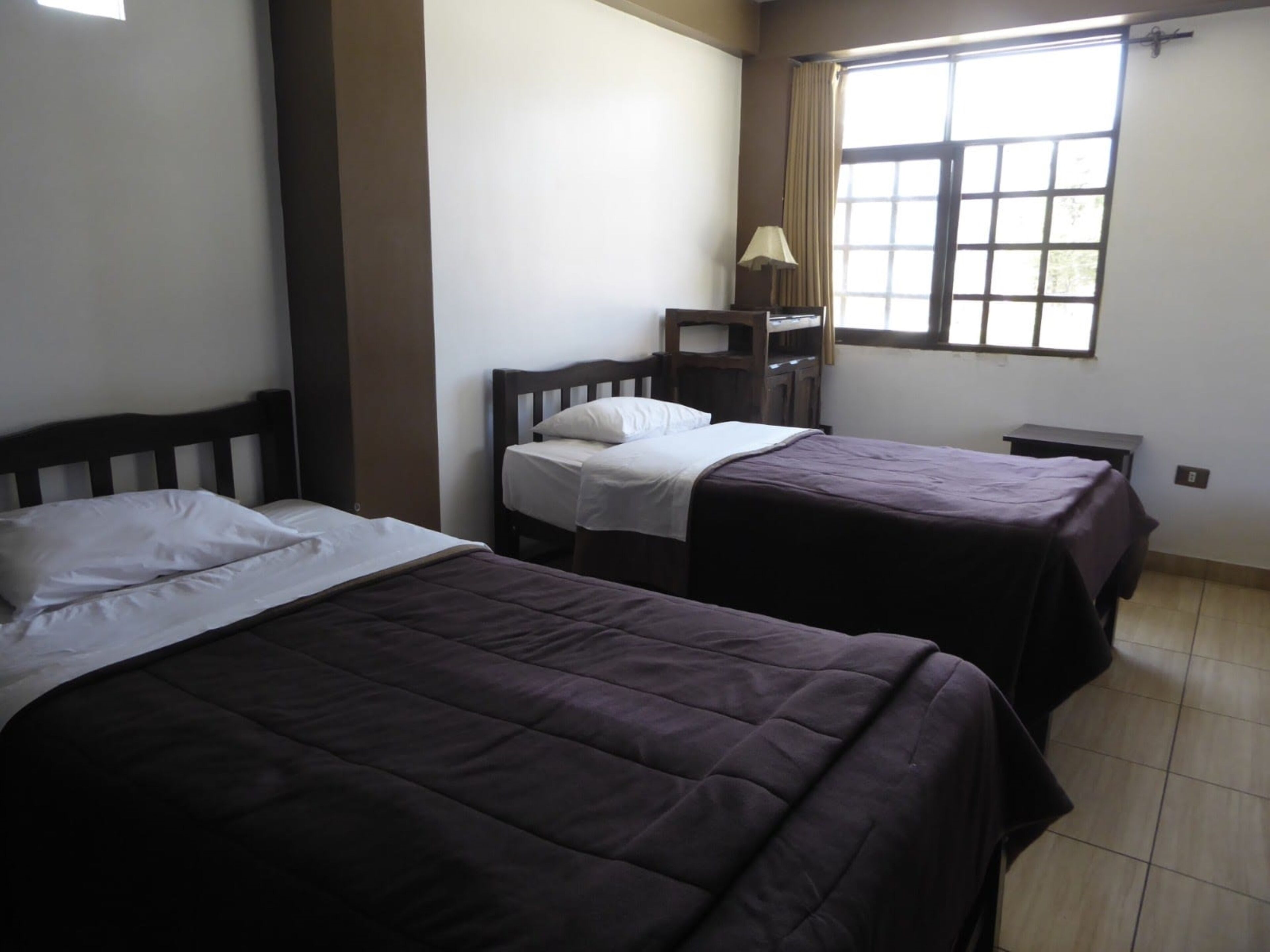 Quarto com duas camas, uma janela com cortinas, uma pequena mesa e um abajur.
