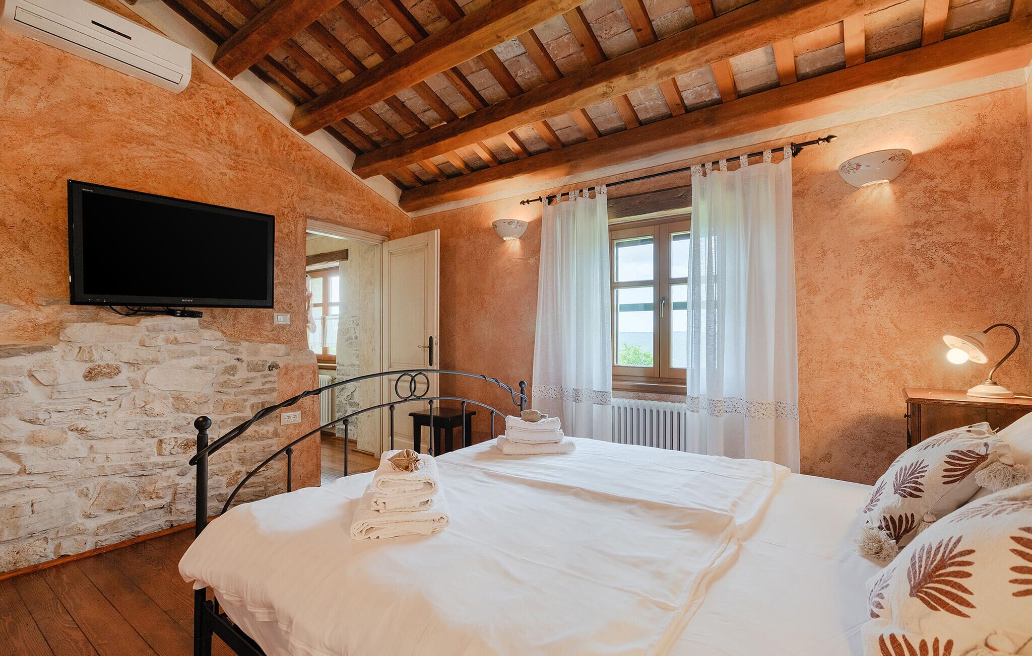 Ein Schlafzimmer mit Steinwand, Holzbalkendecke, einem Bett mit weißer Bettwäsche, einem Fernseher und einem Fenster mit Vorhängen.