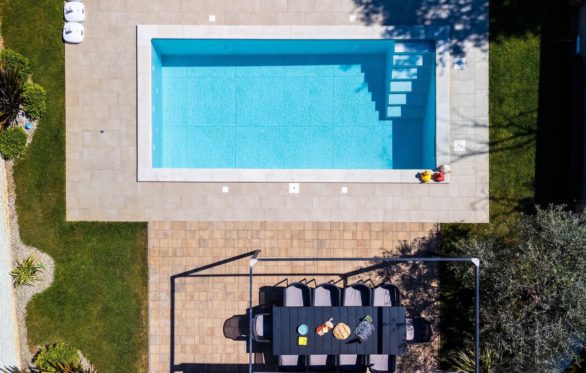 A rectangular swimming pool with a tiled deck surrounding it, a seating area with a table and chairs, and a small garden with plants and a tree.