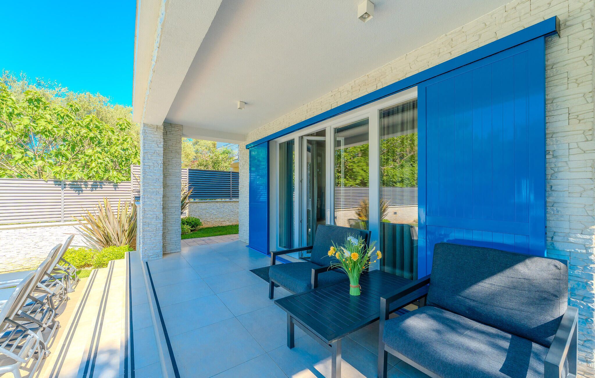 A modern outdoor patio with a dining table and chairs, a glass door leading to a covered area, and a stone wall.