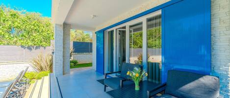 A modern outdoor patio with a dining table and chairs, a glass door leading to a covered area, and a stone wall.
