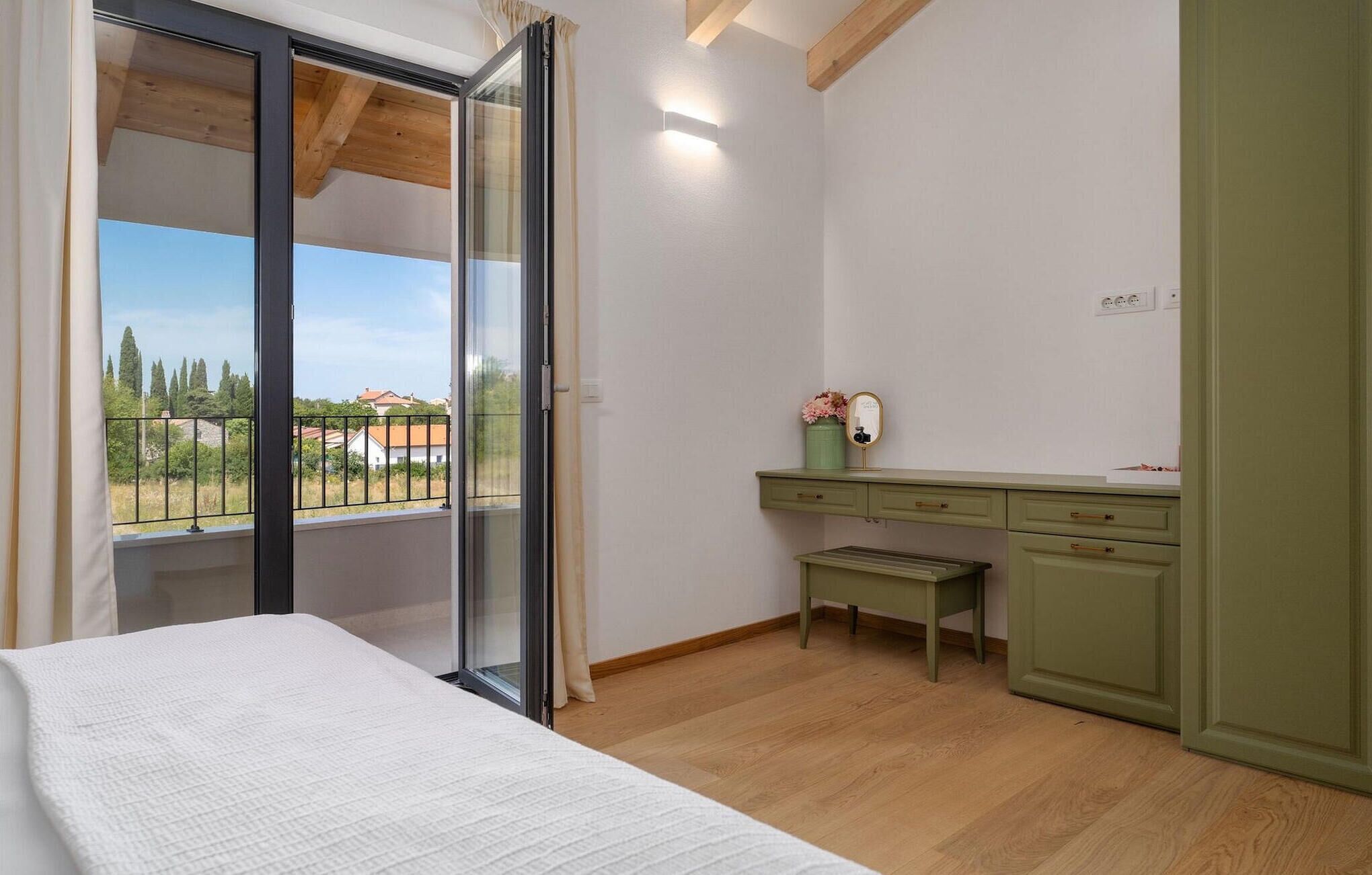 A bedroom with a bed, a desk, a chair, and a balcony with a view of houses and trees.
