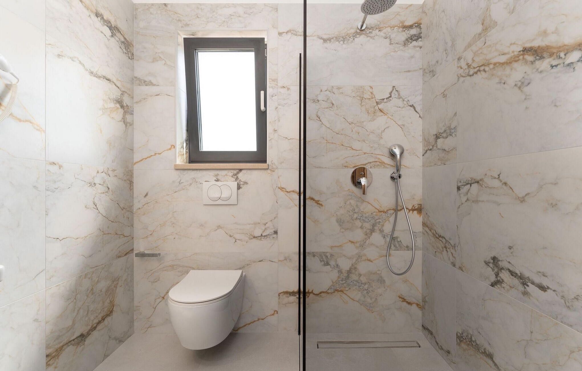 A bathroom with a toilet, a shower with a handheld showerhead, and a window above the toilet.