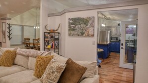 Una sala de estar con un sofá, una estantería y un comedor con sillas. Se ve una cocina a través de una puerta abierta.