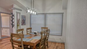 Un comedor con mesa y sillas de madera, una lámpara moderna y ventanas grandes con persianas.