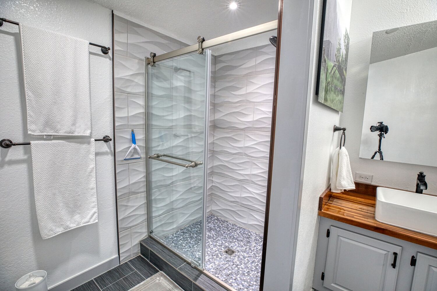 A modern bathroom with a glass shower enclosure, white textured walls, a wooden vanity, and a mirror with a camera on a tripod.