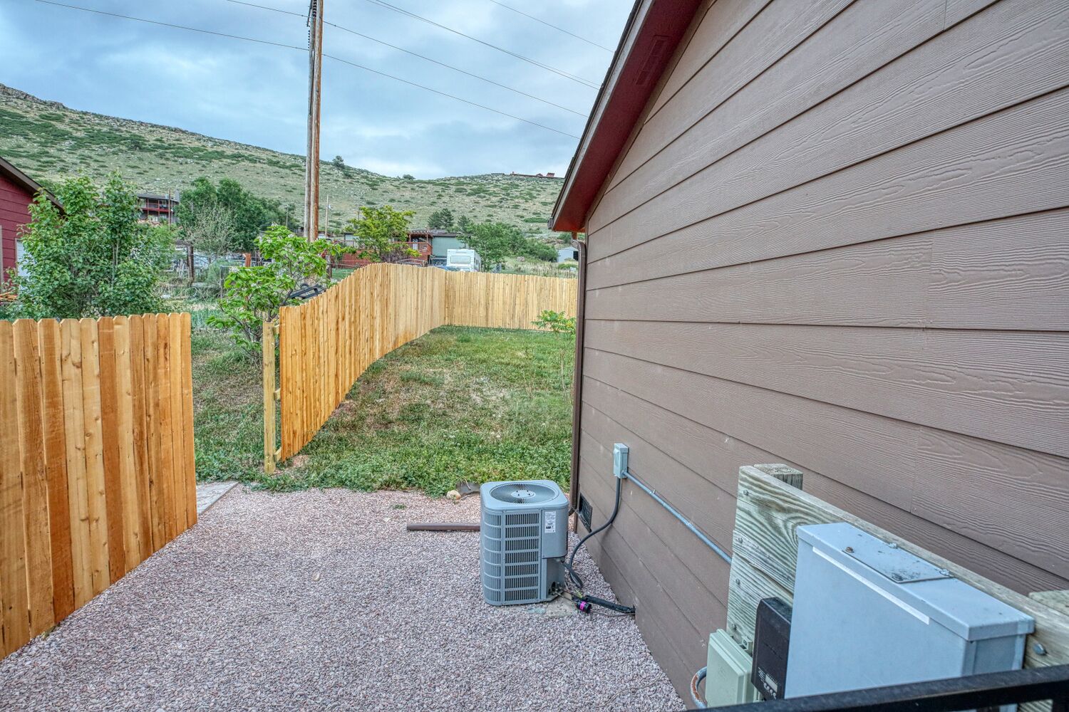A residential backyard with a wooden fence, a gravel patio, and an air conditioning unit mounted on the house.
