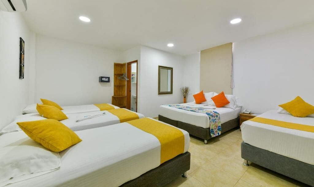 Habitación de hotel con tres camas individuales, cada una con un cojín amarillo decorativo y una colcha blanca. Se observa un armario de madera, una mesita de noche con lámpara y un espejo grande en la pared.