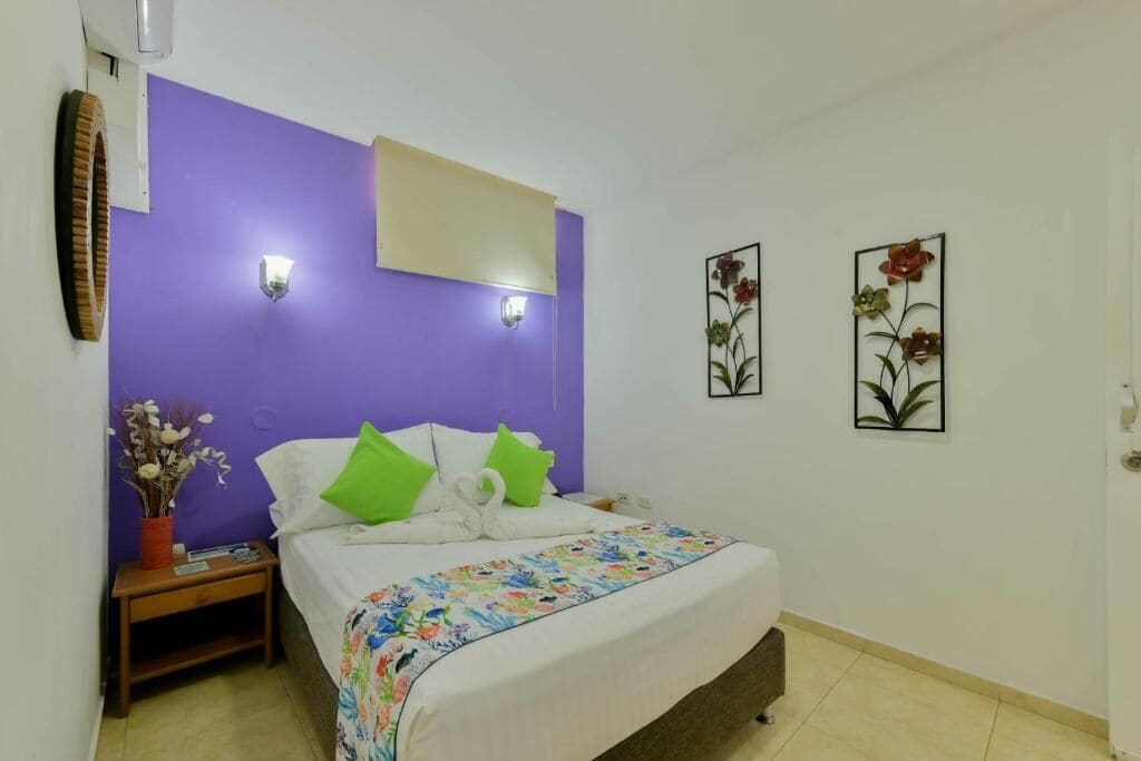 Ein Schlafzimmer mit lila Akzentwand, einem Bett mit weißer Bettwäsche und grünen Kissen, einem Nachttisch mit einer Vase getrockneter Blumen und zwei Wandleuchten.