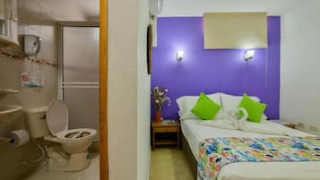 A hotel room with a purple accent wall, a bed with a floral bedspread, a nightstand with a vase of flowers, and a bathroom with a toilet and sink.