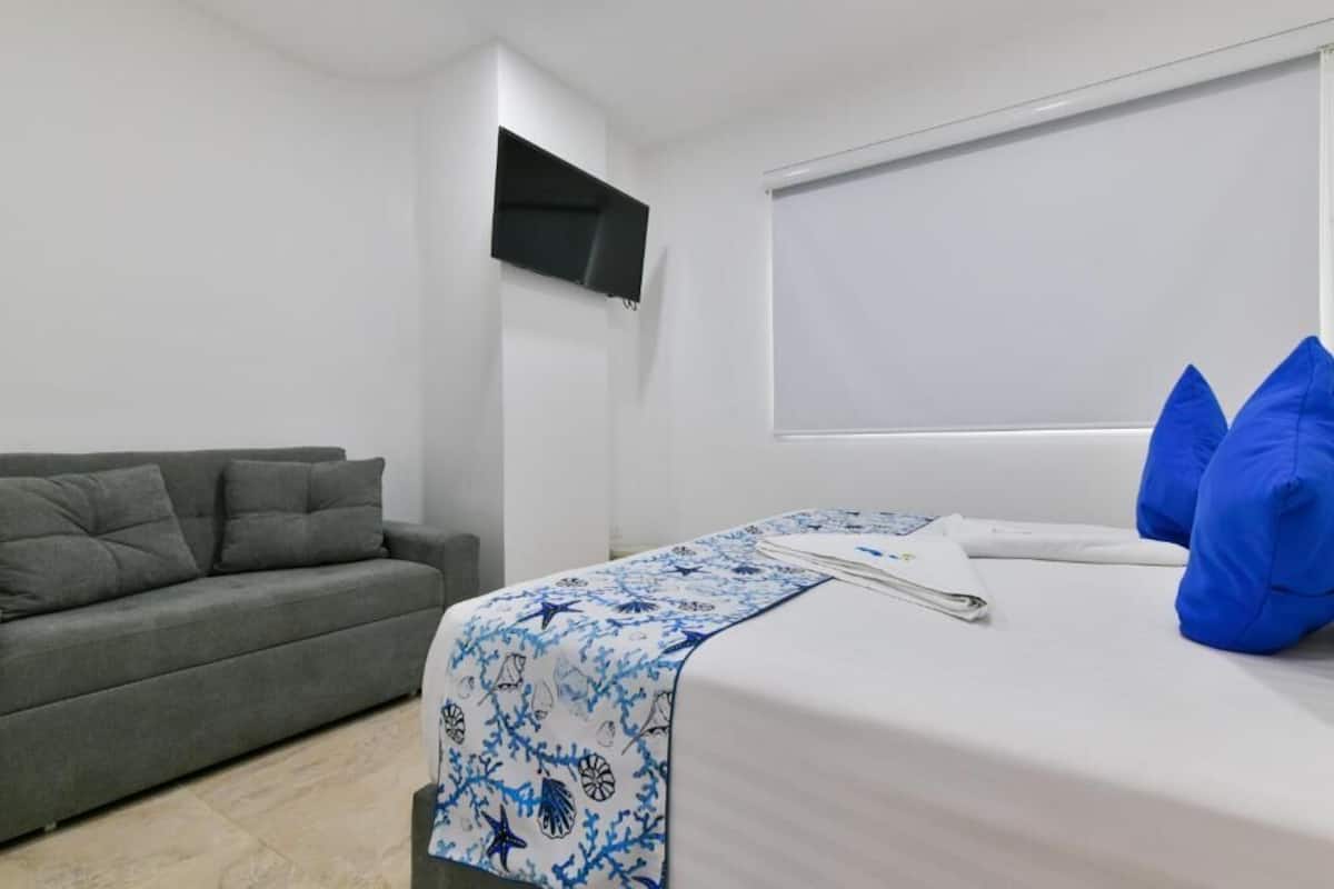 Una habitación de hotel moderna con un sofá gris, una cama con colcha estampada de flores azules y un ventanal con persianas.