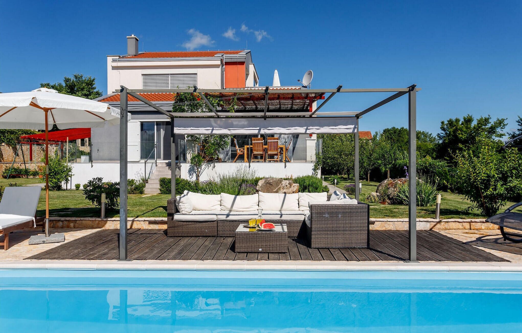 A modern outdoor pool area with a covered seating area, a pool, and a house in the background.