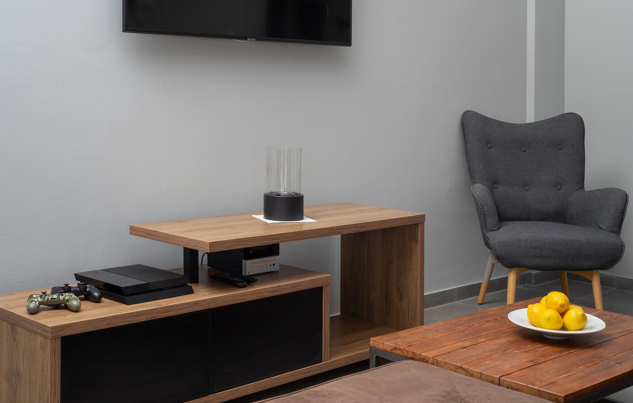 A modern living room with a wooden coffee table, a grey armchair, a TV mounted on the wall, and a small side table with a blender and remote controls.