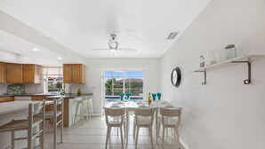 Una cuina moderna amb armaris blancs, una zona de menjador amb cadires blanques i una finestra gran amb vista a una piscina.