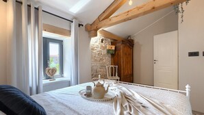 A bedroom with a bed, a window with curtains, a stone wall, a wooden door, and a bedside table with a teapot and cups.
