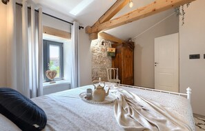 Une chambre à coucher avec un lit, une fenêtre avec des rideaux, un mur en pierre, une porte en bois et une table de chevet sur laquelle se trouvent une théière et des tasses.
