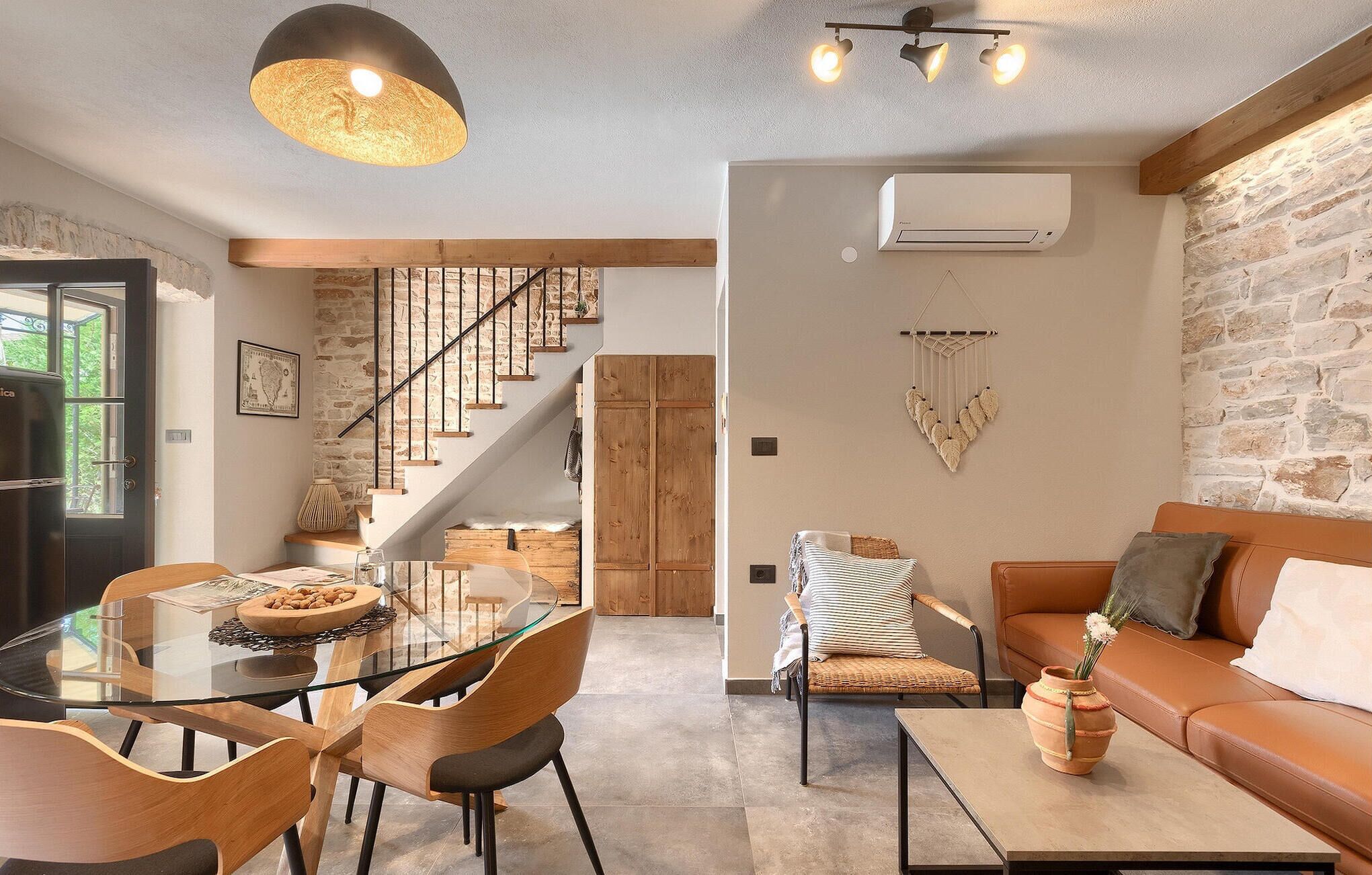 A modern living room with a glass dining table, wooden chairs, a leather sofa, and a staircase with wooden railings.