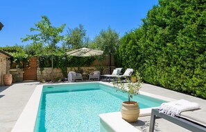 A rectangular swimming pool with a stone wall and a covered patio area with lounge chairs and an umbrella.