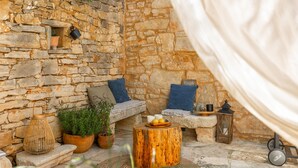 A stone-walled outdoor seating area with a wooden bench, cushions, potted plants, and a round wooden table.