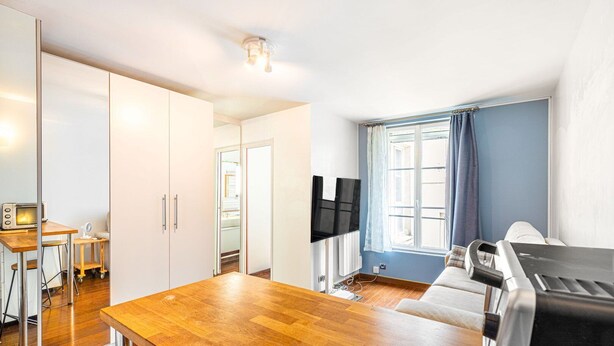 Un espace de vie compact comprenant une table à manger en bois, un téléviseur fixé au mur et une fenêtre avec des rideaux.
