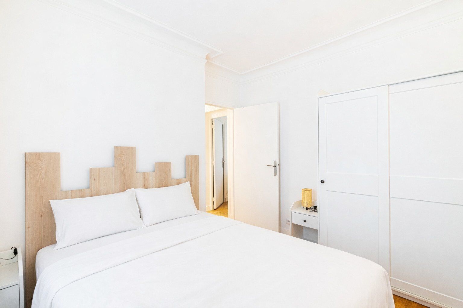 A bedroom with a wooden headboard, a bed with white bedding, a nightstand, a small table with a lamp, and a wardrobe.