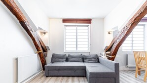 Un dormitorio estilo loft moderno con vigas de madera a la vista, un sofá seccional gris, una silla de madera y una ventana con persianas.