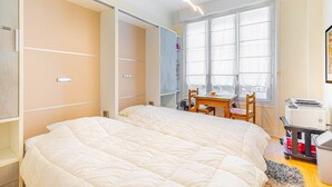 Quarto com cama grande, escrivaninha com cadeira, janela com persianas e impressora sobre um carrinho.