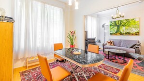 Uma sala de estar moderna com área de jantar, apresentando uma mesa redonda de vidro, cadeiras laranja, um sofá e uma grande janela com cortinas leves.