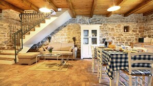 Et rustikt interiør med stenvæg, træloft, trappe, sofa, sofabord, spisebord med stole og et skab.