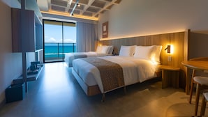 A hotel room with two beds, a wooden headboard, a small wooden table, a chair, a desk, and a large window with a view of the sea. - Ritz Barra de São Miguel (Barra de São Miguel)