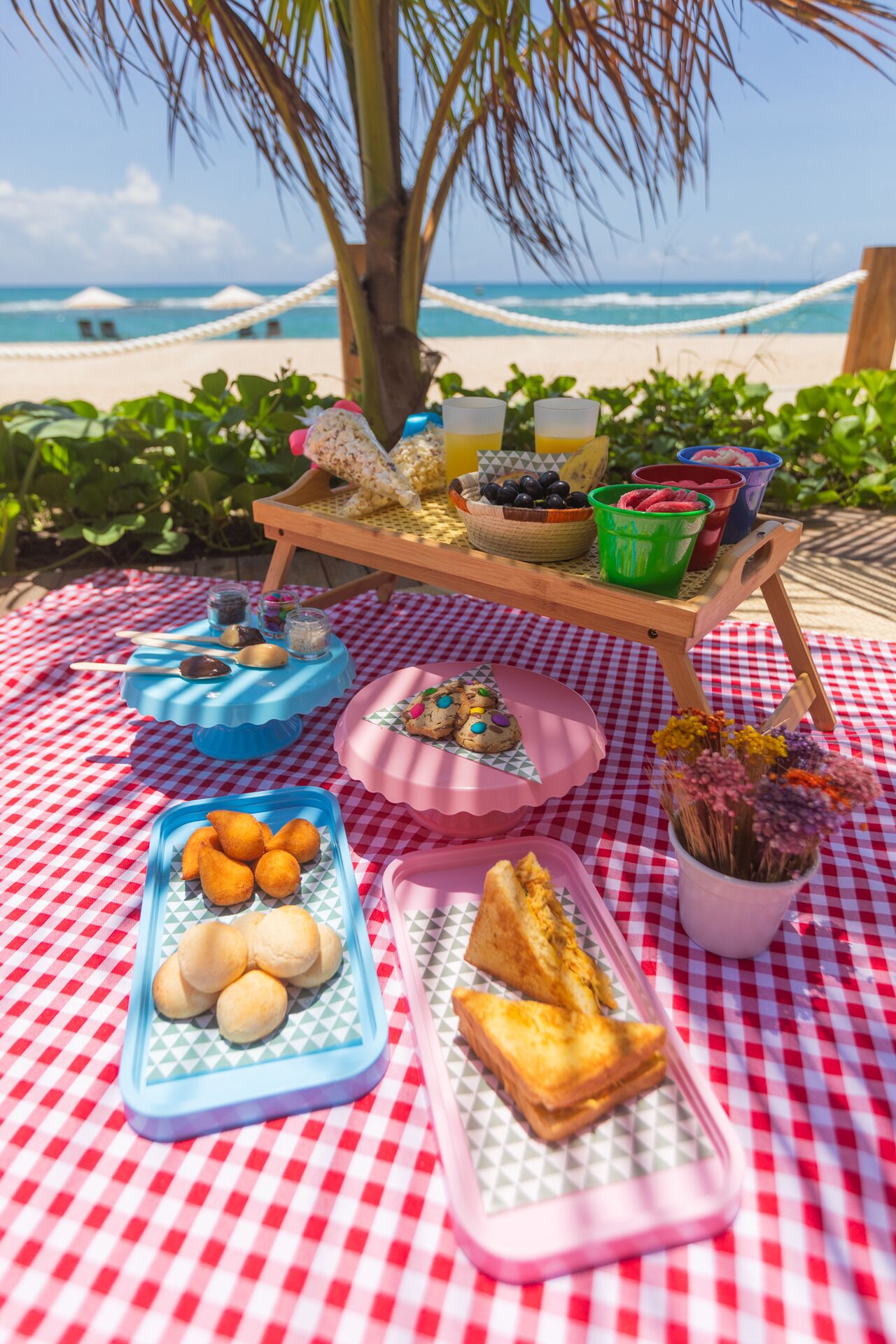 Ein Picknick am Strand mit karierter Tischdecke, einem Holztablett mit Snacks und Getränken und Blick aufs Meer im Hintergrund.