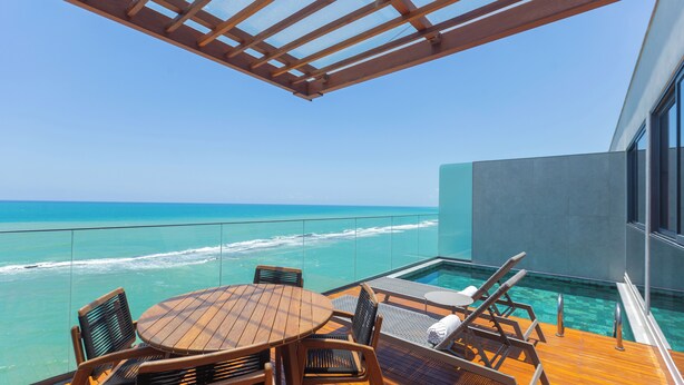 A wooden deck with a table and chairs, overlooking a clear blue ocean and a swimming pool. - Ritz Barra de São Miguel (Barra de São Miguel)