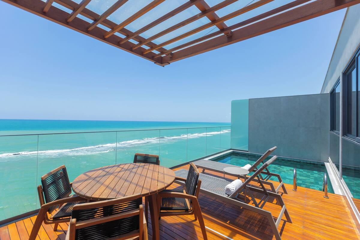 Une terrasse en bois avec une table et des chaises, offrant une vue sur un océan d’un bleu limpide et une piscine.