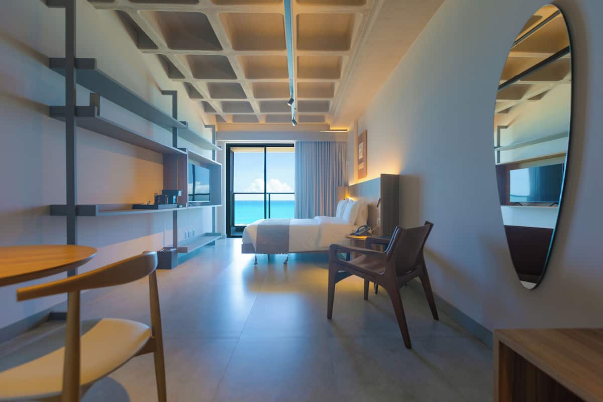 Une chambre d’hôtel moderne dotée d’un grand lit, d’un bureau, d’une chaise, d’un miroir et offrant une vue sur l’océan.