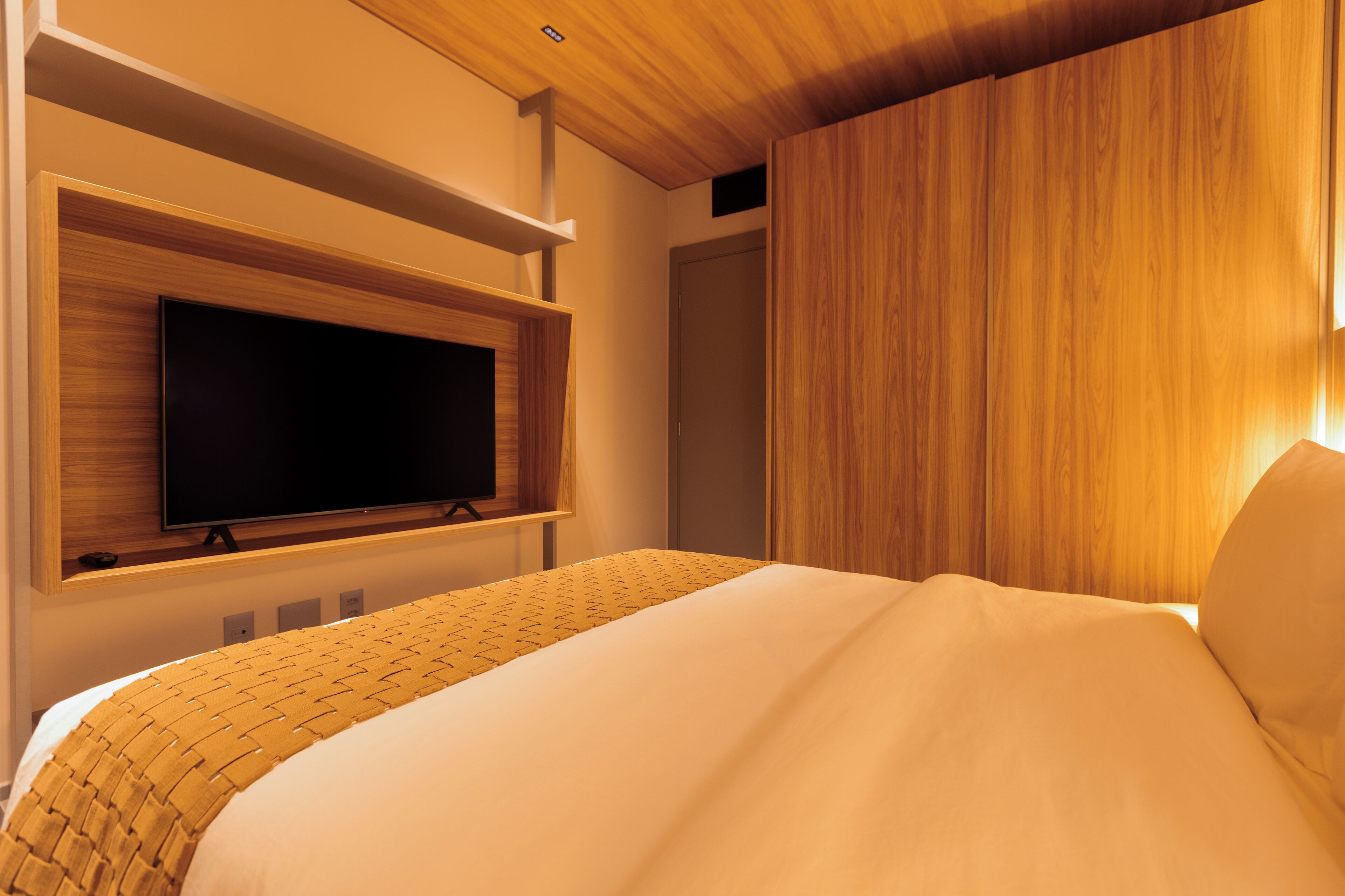 A modern hotel room with a large bed, a mounted flat-screen TV, wooden paneling, and a built-in shelf.