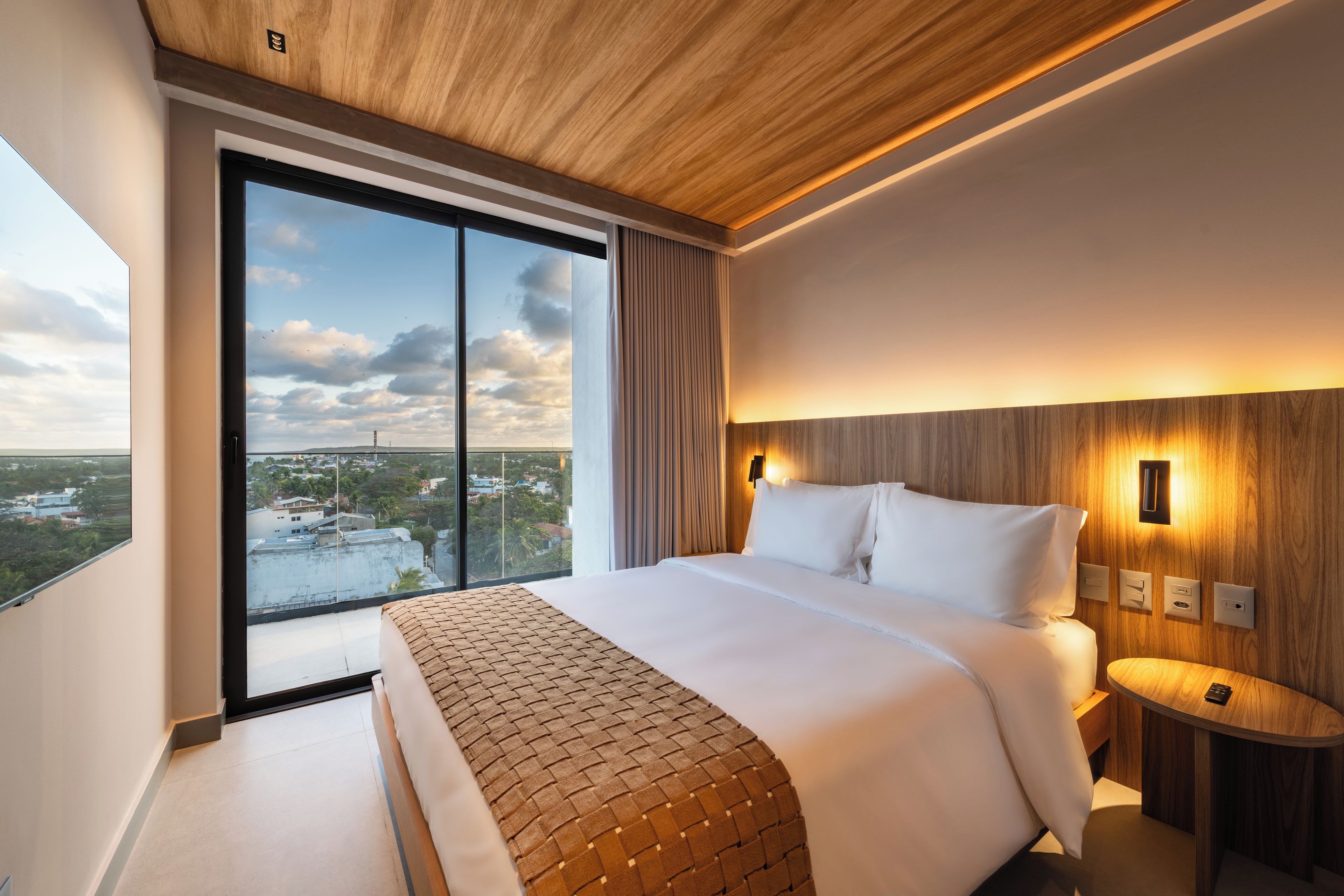 Ett modernt hotellrum med en stor säng, ett nattduksbord, en väggmonterad lampa och utsikt över staden genom ett stort fönster.