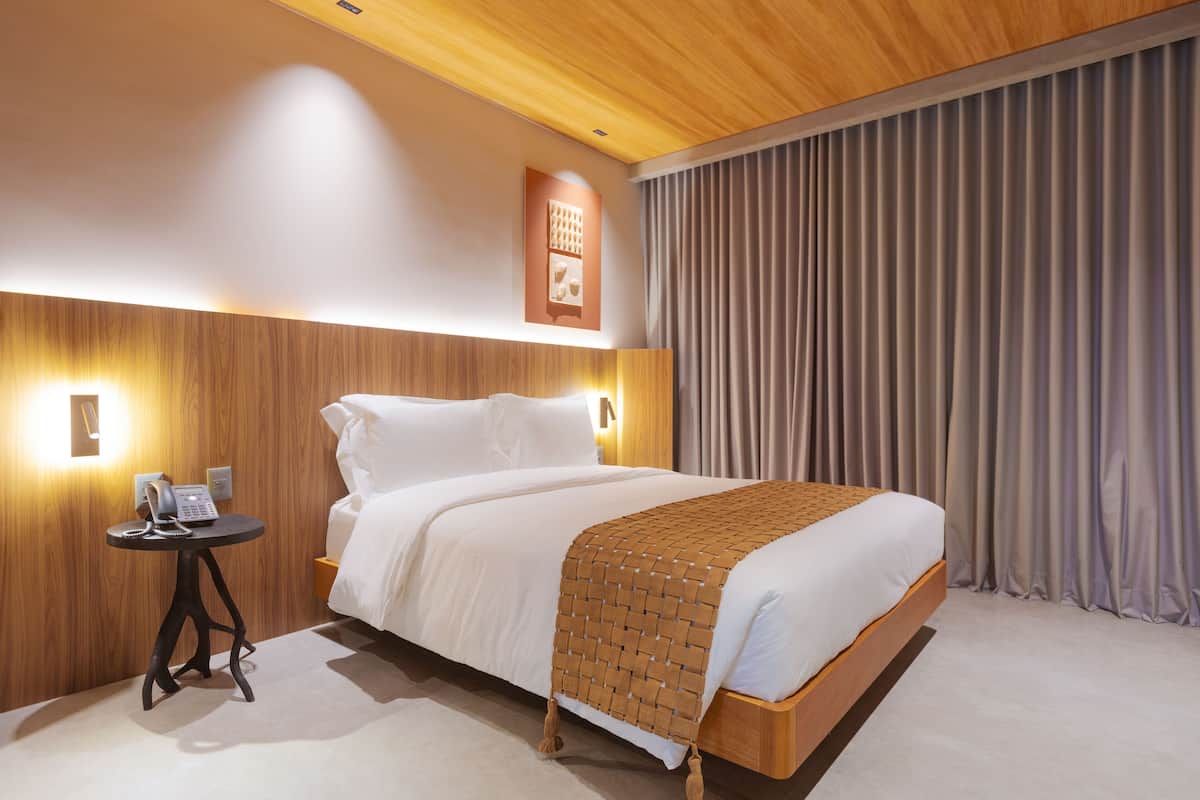 Une chambre d’hôtel moderne avec une tête de lit en bois, une table de chevet avec un téléphone et un grand lit recouvert d’une couverture tissée.