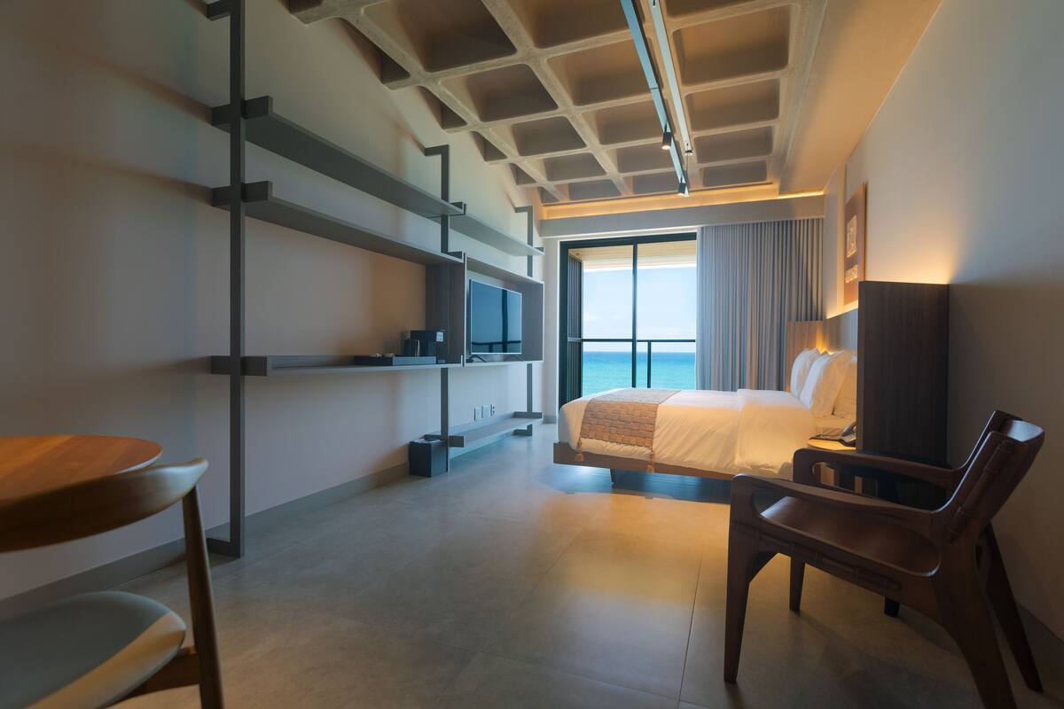 Une chambre d’hôtel moderne équipée d’un lit, d’un bureau, d’une chaise et d’une grande fenêtre donnant sur l’océan.