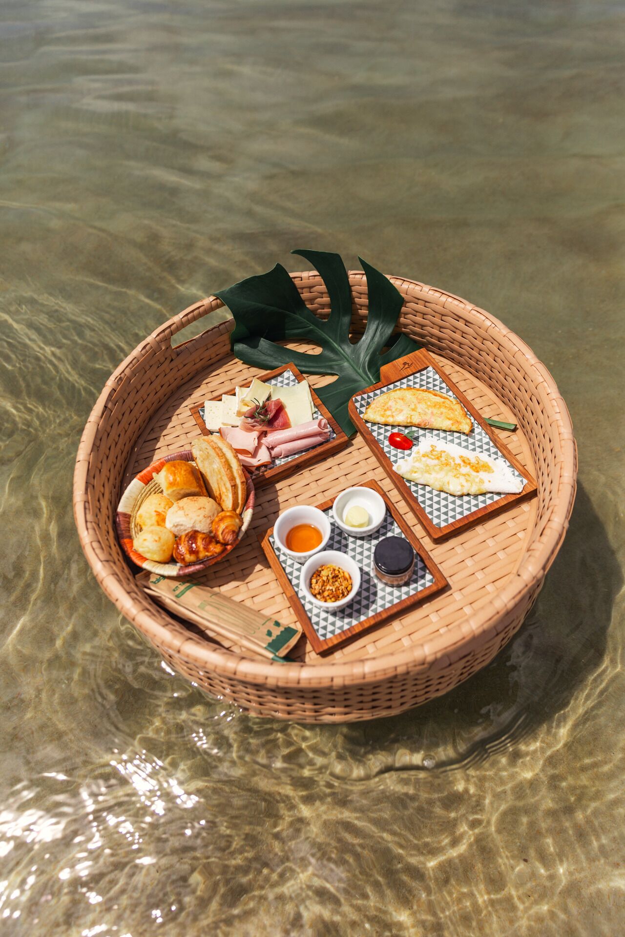 A wicker basket filled with various food items, including bread, cheese, and condiments, placed on a clear surface.
