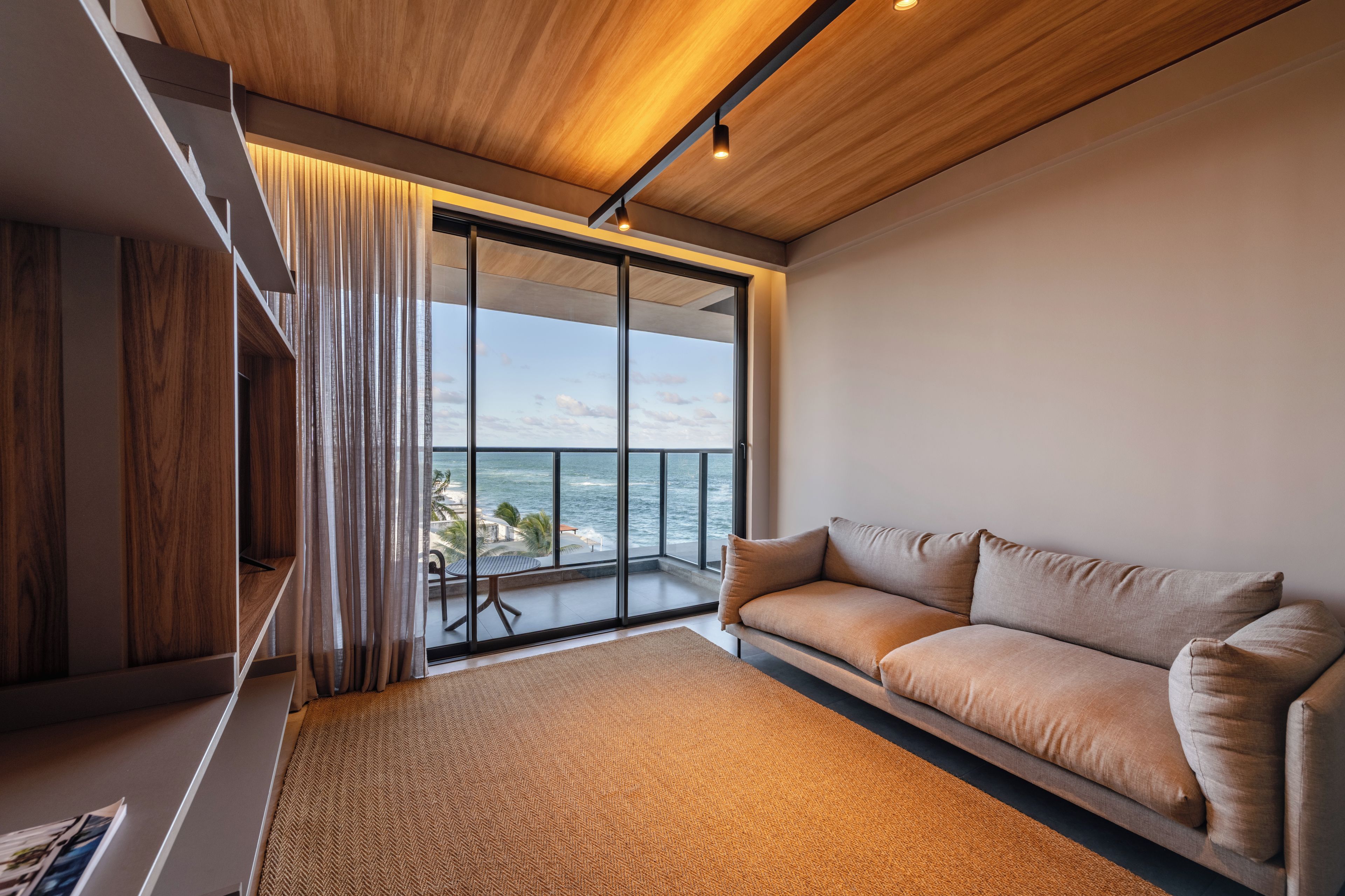 A modern living room with a large window overlooking the sea, a sofa, and a wooden ceiling.