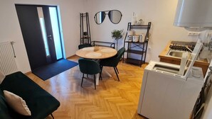 Un espai vital compacte amb un sofà, una taula de dinar amb cadires, una zona de cuina amb fogó i rentapeus, i una porta que dona a un balcó.