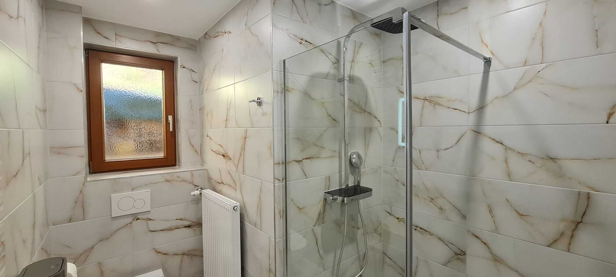 A bathroom with a glass shower enclosure, marble tiles, a window with a view, and a radiator.