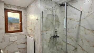 A bathroom with a glass shower enclosure, marble tiles, a window with a view, and a radiator.