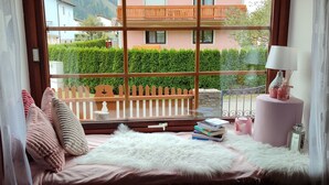 Habitació acollidora amb una finestra gran, un llit confortable amb almoades, una taula de nit amb llibres i una làmpada, i vista a un balcó i vegetació a l'exterior.