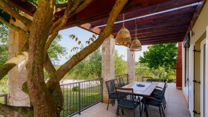 A covered patio with a table and chairs, a hanging light fixture, and a view of greenery and trees.